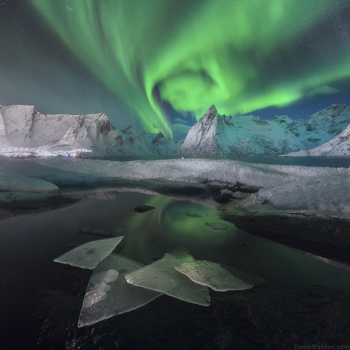 Brilliant green aurora borealis swirls above dramatic Arctic peaks, its vivid light reflecting in the dark waters where geometric ice formations create a natural sculpture garden