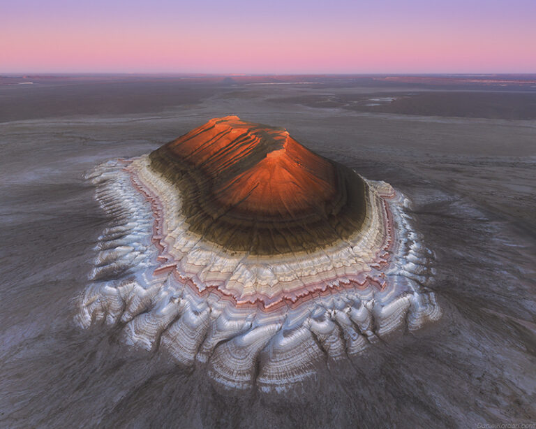 Bokty Mountain rises 165 meters above the vast plains of West Kazakhstan, its vibrant layered rock formations resembling a giant natural cake. The mountain's striking colors reach their most magical intensity during sunrise and sunset, when they glow with fiery light and cast an ethereal glow across the landscape.