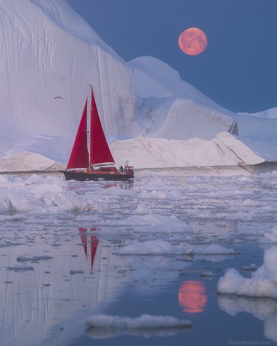 Red-sailed schooner navigates Antarctic waters beneath the luminous glow of a full moon.