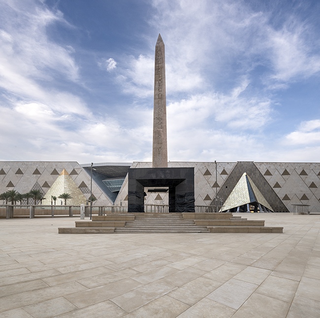 Suspended obelisk in a wide plaza with the museum walls and pyramids beyond.