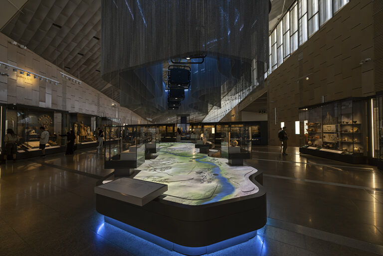 Long gallery with illuminated Nile relief table and hanging mesh above display cases.
