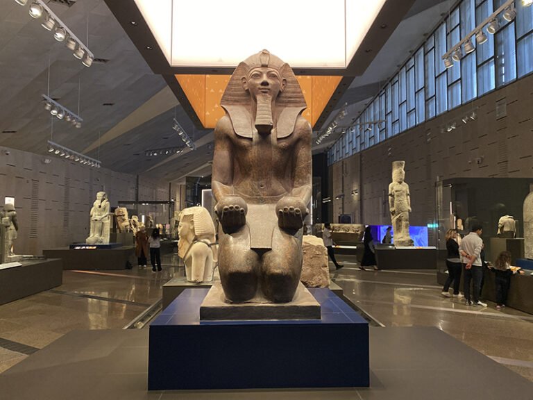 Seated statue of Queen Hatshepsut surrounded by vitrines and visitors.