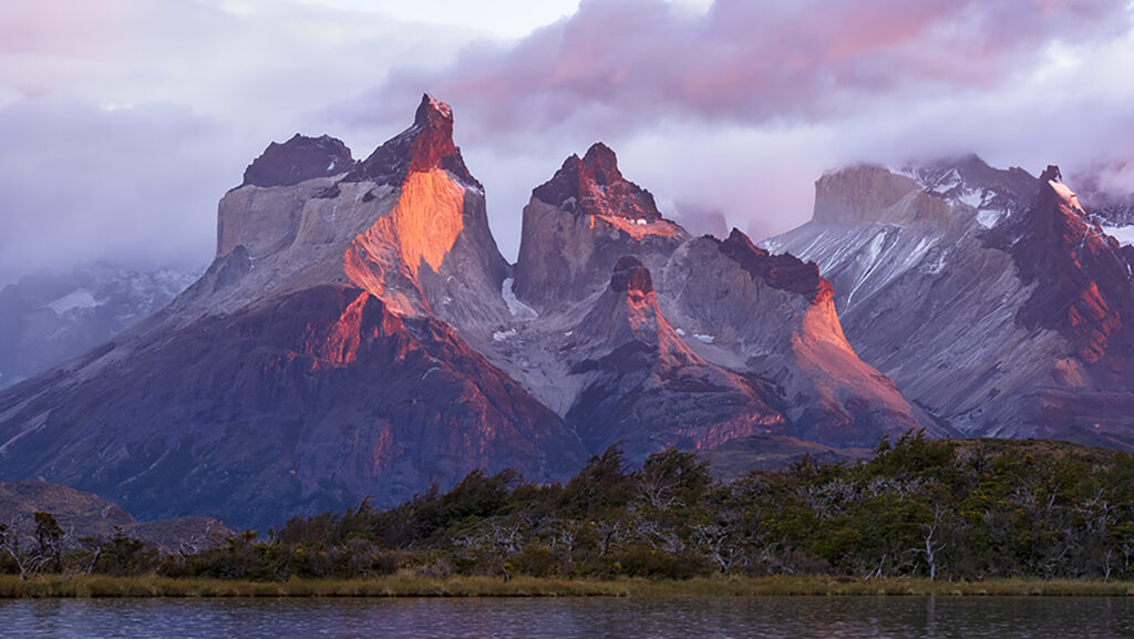 The peaks of Torres del Paine catch the first light of sunrise, reflected in the still water below.