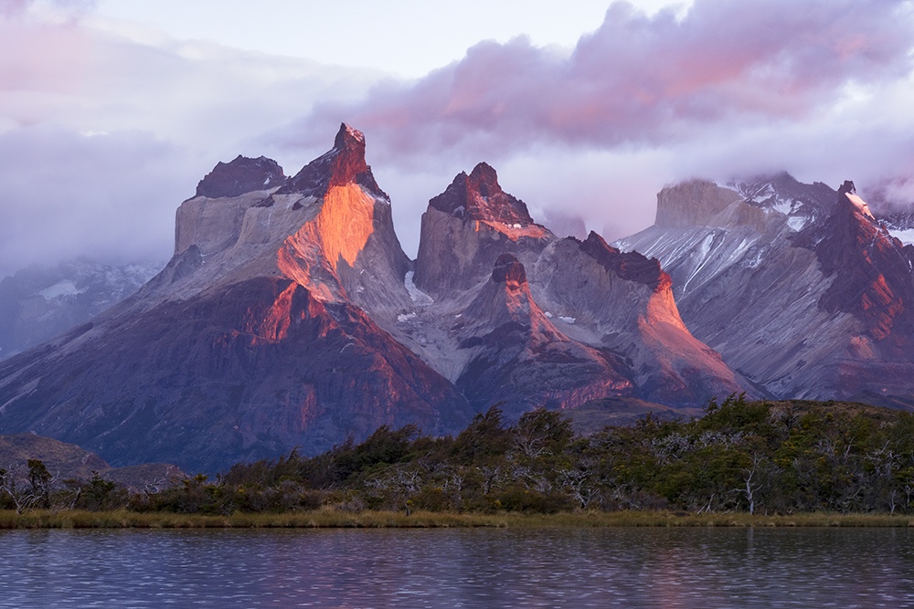 The peaks of Torres del Paine catch the first light of sunrise, reflected in the still water below.